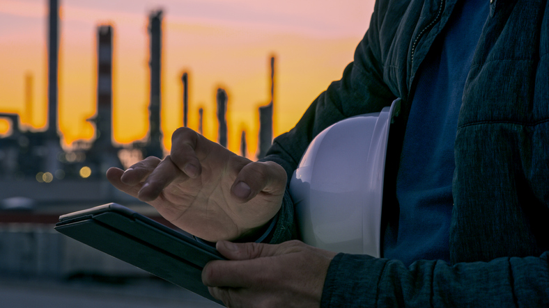 Close-up shot of the hands an engineer with a white hardhat using a tablet with an oil refinery visible in the background during sunset.
